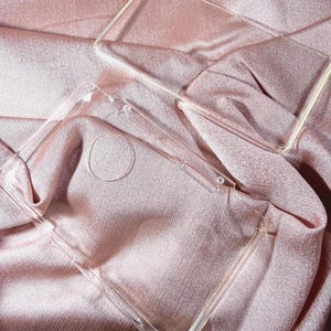 Puede incluir: Dos fundas transparentes para teléfono se muestran sobre una tela de satén rosa. Las fundas son rectangulares con recortes para la cámara y los botones. La tela tiene una textura suave y fluida.