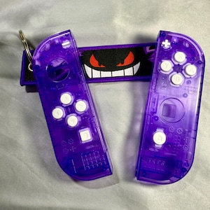 May include: A pair of transparent purple game controllers with white buttons and a matching wrist strap. The strap is black with a smiling ghost face design in red and white. A silver key ring is attached to one controller.