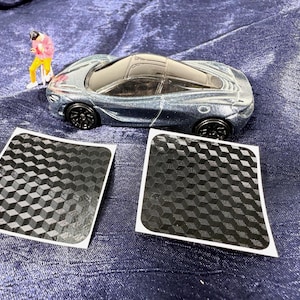 May include: A miniature blue-gray sports car with black wheels and a black roof sits on a blue surface. Two black and white patterned decals are in front of the car. A small figurine stands near the car.