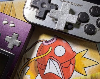 May include: Close-up of two handheld gaming consoles, one purple and one silver, with directional pads and buttons. A gray directional pad is placed on top of the silver console. The background features a colorful cartoon fish design.