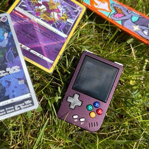 May include: A purple handheld gaming device with a screen, directional pad, and colored buttons, alongside several Pokémon trading cards and a Charizard keychain. The cards feature Radiant Alakazam and other Pokémon characters.