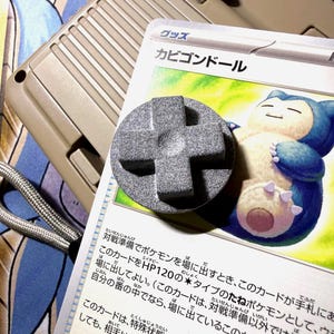 May include: A gray, cross-shaped button sits atop a Pokémon trading card featuring a blue and white Snorlax illustration. A tan handheld gaming device is in the background, with a gray lanyard.