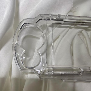 Może przedstawiać: Przezroczyste, plastikowe etui ochronne na przenośne urządzenie do gier. Etui ma wyprofilowaną konstrukcję z otworami na przyciski i ekran oraz jest wykonane z przezroczystego, twardego plastiku.