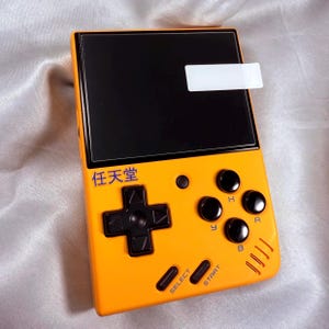 May include: An orange handheld gaming console with a black screen and black buttons. The console has a directional pad, action buttons, and text that reads "SELECT" and "START". The top left corner has Japanese text.