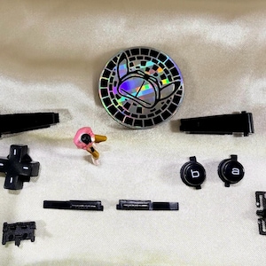May include: An assortment of black video game controller parts and accessories, including buttons, directional pads, and a decorative coin with a holographic design. A small, pink figurine is also present.