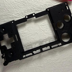 May include: A black, rectangular, textured plastic frame component featuring a large central rectangular cutout, a cross-shaped opening on the left side, and three circular apertures on the right side, with various small mounting holes.