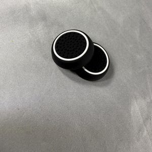 May include: Two black controller thumb grips with a white rim and a textured surface. The grips are circular and designed to enhance gaming control. The grips are shown on a gray surface.