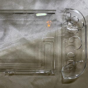May include: Transparent plastic protective case for a gaming console, comprising a rectangular main section and a curved side piece with button apertures. The case is designed to safeguard the console from scratches and damage.