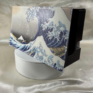 May include: A black electronic device with a decorative cover featuring the iconic "Great Wave off Kanagawa" artwork. The cover displays a blue and white wave design with a mountain in the background. The device sits on a white, circular base.