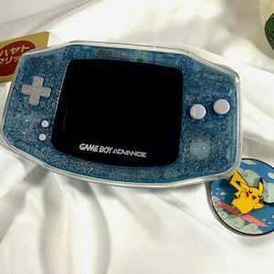 Könnte beinhalten: Ein blaues Game Boy Advance Handheld-Spielkonsolen mit einer glitzernden, transparenten Hülle. Die Konsole verfügt über einen schwarzen Bildschirm, ein Steuerkreuz und A- und B-Tasten. Der Text "GAME BOY ADVANCE" ist auf der Vorderseite aufgedruckt.