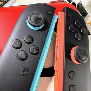 May include: Close-up of two black Nintendo Switch Joy-Con controllers. One controller has a light blue side, and the other has an orange side. Each controller has black buttons and joysticks with colored accents.