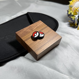 May include: Two red and white Pokeball-themed accessories rest on a small, square, wooden block. A black case is in the background, and a small figurine is visible in the upper right corner. The accessories are likely for a gaming device.