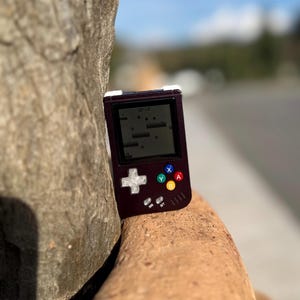 May include: A handheld retro gaming console in a dark burgundy case. The screen displays a classic video game. The console has a directional pad, colored action buttons, and a white cross-shaped button. The device is resting on a rough, brown surface.