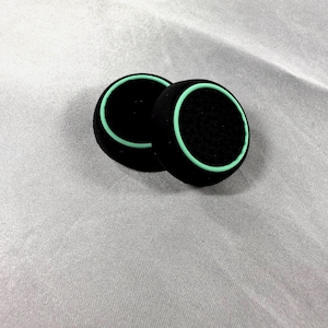 May include: Two black foam ear pads with a light green trim. The ear pads are circular and have a textured surface. The pads are designed for headphones or headsets.