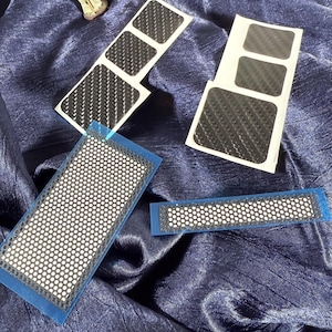 May include: Various black carbon fibre-style stickers with a textured pattern are shown on a dark blue fabric. Some stickers are rectangular, while others have a perforated design with a blue border. A small Yoda figurine is also present.