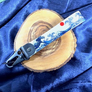 May include: A key fob strap with a Japanese wave pattern in blue and white, and a red sun. It has a black metal clasp and leather backing. The strap is displayed on a wooden slice.