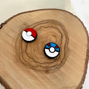 以下が含まれることがあります： 白を基調とした丸いカラフルなチャームが2つ。1つは赤と白、もう1つは青、白、赤。自然な木目模様の木製の表面に配置されています。