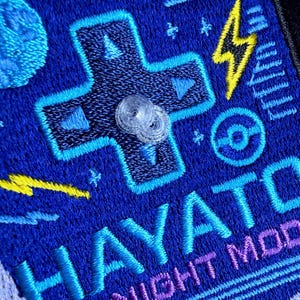 May include: A blue embroidered patch featuring a light blue D-pad, a yellow lightning bolt, a circular icon, and the text HAYATO NIGHT MODE in light blue and purple thread. A small, clear, circular plastic component is attached to the centre.