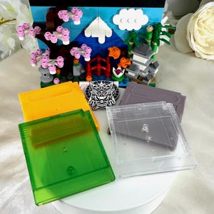 May include: A collection of Game Boy cartridges in yellow, green, clear, and gray. A small diorama of a Japanese scene with a red gate, cherry blossoms, and a pagoda is in the background. A black and white circular emblem is also present.