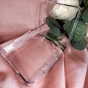 Può includere: Una custodia rettangolare trasparente in acrilico con coperchio incernierato, aperta per rivelare l'interno. La custodia è su una superficie di raso rosa, con fiori bianchi e foglie verdi sullo sfondo. La custodia ha una finitura liscia e lucida.