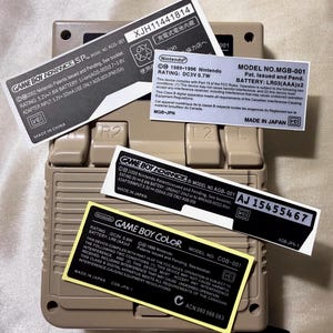 Könnte beinhalten: Eine beige Game Boy Advance SP-Konsole mit mehreren Aufklebern. Die Aufkleber enthalten Text wie "GAME BOY ADVANCE SP" und "MADE IN CHINA". Die Konsole hat Knöpfe und einen Bildschirm.