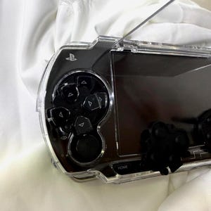May include: Transparent protective case for a handheld gaming console. The case is clear, showing the console's buttons and screen. The console's buttons are black, and the case has a sleek, modern look. The word "HOME" is visible on the console.