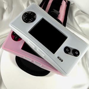 May include: Two handheld gaming consoles, one white and one pink, are stacked on top of each other. The white console has a black screen and black buttons. The pink console is visible beneath the white one. A black and pink carrying case is in the background.