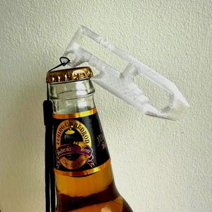 May include: A clear plastic bottle opener is attached to a bottle of butterscotch beer. The bottle has a black label with gold lettering and a gold cap. The opener is white and has a handle.
