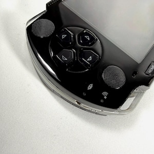 May include: A black handheld games console with a glossy finish. The device has a directional pad, action buttons, and a screen. The console has a silver trim and a textured grip area. The "HOME" button is visible.