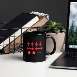 May include: A black ceramic mug with a red controller graphic. The graphic includes directional arrows and the letters A and B. The mug is on a white surface with a laptop, a plant, and a wire basket in the background.