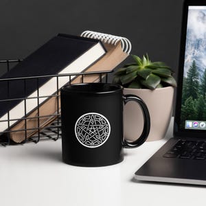May include: A black ceramic mug with a white Celtic knot design. The mug is on a white surface, with a laptop, a succulent plant, and a wire basket with notebooks in the background. The mug has a glossy finish.