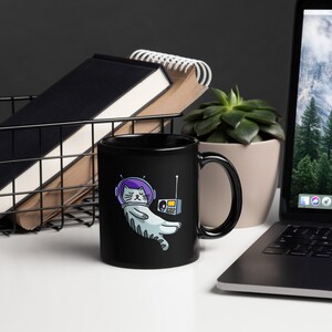 May include: Black ceramic mug featuring a cartoon cat in a space helmet, holding a retro radio. The cat is light gray with purple accents. The mug is on a white surface, with a laptop, plant, and books in the background.