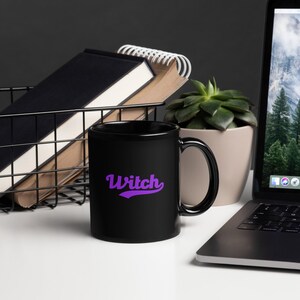 May include: A black ceramic mug with the word "Witch" in purple script. The mug is on a white surface, next to a laptop, a succulent plant in a beige pot, and a wire basket with books.