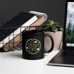 May include: Black ceramic mug featuring a pentagram design with vines and white flowers. The pentagram is encircled by a gray border. The mug is on a white surface, with a laptop, books, and a plant in the background.