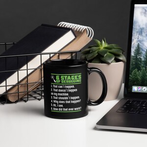 May include: A black ceramic mug with the text "6 Stages of Debugging" in green and white. The text lists common debugging phrases. The mug is on a white surface near a laptop and a succulent plant.