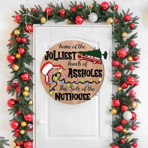 May include: A round wooden Christmas sign with the text "home of the jolliest bunch of assholes this side of the nuthouse". The sign features a Santa's hand, a car with a Christmas tree, and Christmas lights. The sign is surrounded by a garland of red and gold ornaments.