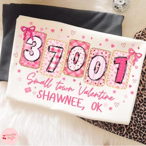 Puede incluir: Una camiseta blanca con un diseño de San Valentín. Presenta los números "37001" en rosa, adornados con corazones y lazos. El texto "Small town Valentine SHAWNEE, OK" está impreso debajo.