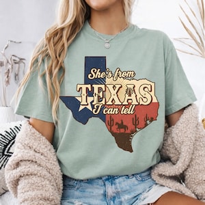 Puede incluir: Camiseta verde claro con un gr&aacute;fico en forma de Texas. El gr&aacute;fico incluye el texto "She's from TEXAS I can tell", una estrella, cactus y la silueta de una persona a caballo. El dise&ntilde;o utiliza colores azul, rojo y beige.
