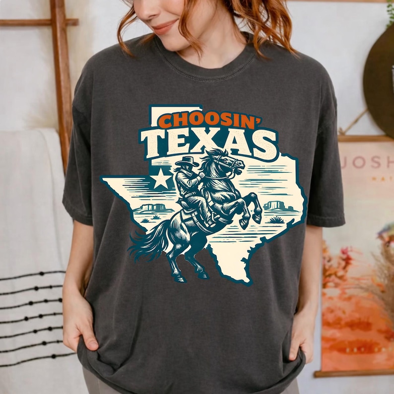 Puede incluir: Camiseta gris oscuro con un gr&aacute;fico de un vaquero montando un caballo encabritado dentro del contorno de Texas. El texto "CHOOSIN' TEXAS" est&aacute; encima del gr&aacute;fico. El dise&ntilde;o utiliza colores azul y crema.