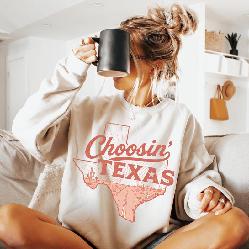 Puede incluir: Una sudadera crema con el texto "Choosin' TEXAS" en naranja-rojo, con un contorno de Texas con un dise&ntilde;o de carretera y cactus. La persona sostiene una taza negra.