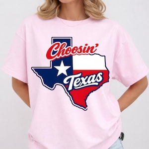 Puede incluir: Una camiseta rosa claro con un gr&aacute;fico del estado de Texas en azul marino, blanco y rojo. Las palabras "Choosin' Texas" est&aacute;n impresas en escritura roja y blanca sobre el contorno de Texas. La camiseta es de estilo casual.