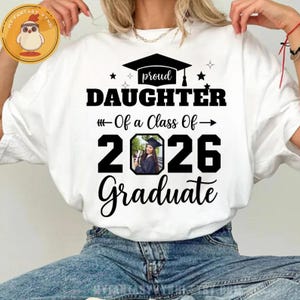 May include: White t-shirt with a graduation-themed design. The shirt features the text "proud DAUGHTER Of a Class Of 2026 Graduate" in black, with a photo of a graduate. A graduation cap and stars are also included.