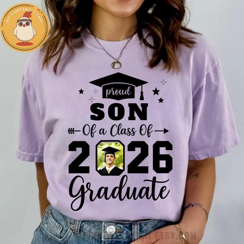 May include: A light purple t-shirt with the text "proud SON of a Class Of 2026 Graduate". The design includes a graduation cap, stars, and a photo of a graduate. The shirt is a comfortable, casual style.