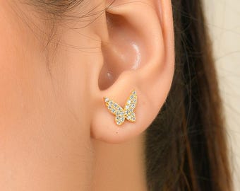 May include: A close-up shot of a person's face and ear, showcasing a gold butterfly earring. The earring is encrusted with small, sparkling stones. The butterfly design is delicate and detailed, adding a touch of elegance to the jewelry.