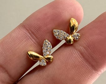 May include: A pair of gold-colored butterfly stud earrings. Each earring features a butterfly design, with one half of the wings encrusted with small, clear gemstones. The earrings are displayed on a finger.