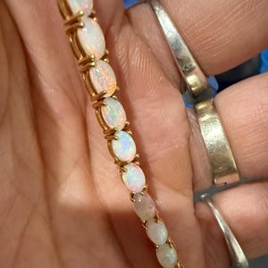 Könnte beinhalten: Ein goldenes Armband mit ovalen Opalen. Die Opale sind in einer Goldkette gefasst und bilden ein zartes und elegantes Schmuckstück. Die Opale haben eine Reihe von Farben, darunter Weiß, Rosa und Orange.