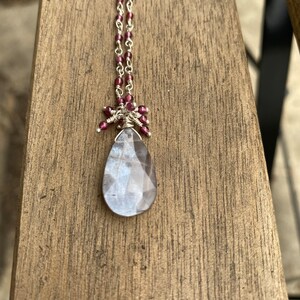 Può includere: Una collana a catena d'argento con un ciondolo a forma di lacrima in cristallo trasparente. Il ciondolo è circondato da piccole perline rosso scuro.