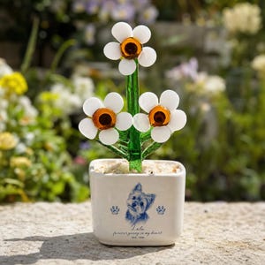May include: A glass flower sculpture with three white petals and amber centers on a green stem, set in a square white ceramic pot. The pot features a blue illustration of a dog, two paw prints, and the text Lulu forever young in my heart 2013-2024.
