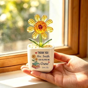 Può includere: Un piccolo vaso quadrato bianco contenente un girasole in vetro giallo con foglie verdi e piccoli sassolini bianchi. Il vaso presenta il testo stampato: THANK YOU Mrs. Smith FOR HELPING ME Grow Love, Lily, con un disegno di un girasole e libri.