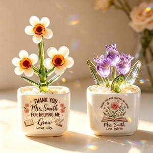 以下が含まれることがあります： 白い正方形のセラミックポットに入った2つのガラス製フラワーアレンジメント。左のポットには白と琥珀色のガラスの花があり、Thank You Mrs. Smith For Helping Me Grow Love, Lilyという文字が書かれています。右のポットには紫色のガラスのチューリップがあり、Thank You For Helping Me Grow Mrs. Smith Love, Oliverという文字が書かれています。